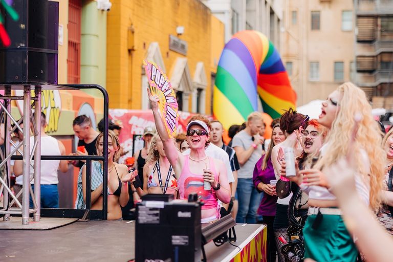 Lively crowd dancing outdoors at the Poppin Out LGBTQ+ festival with a rainbow arch, drag performer, and fan.